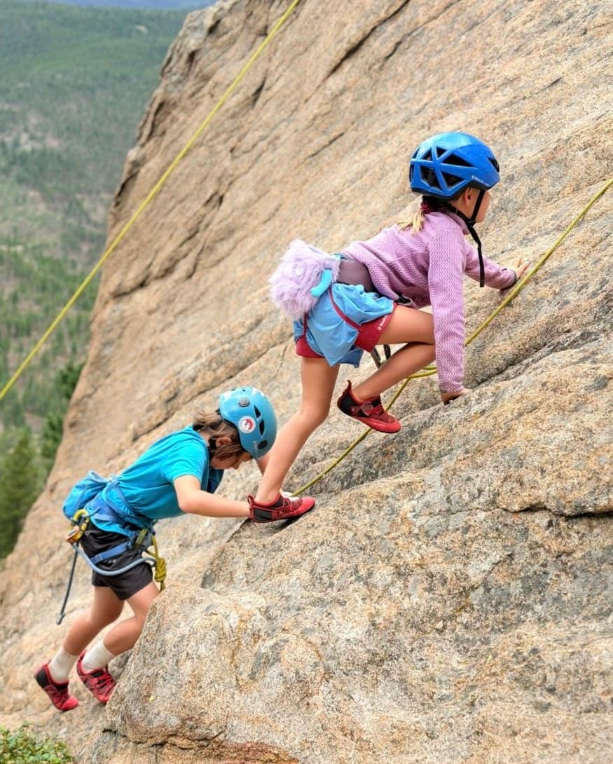 Two kids in helmets climbing a granite slab above a forested valley